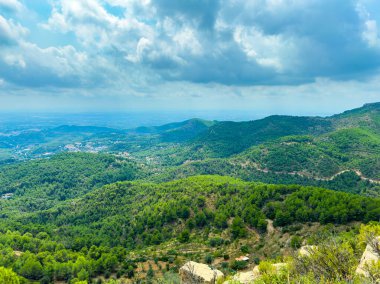 Bulutlu bir gökyüzü tarafından çerçevelenmiş yemyeşil dağlar uzaklığa kadar uzanıyor. Manzara kayan tepeleri ve vadileri gösteriyor, barışçıl, doğal bir ortam yaratıyor..