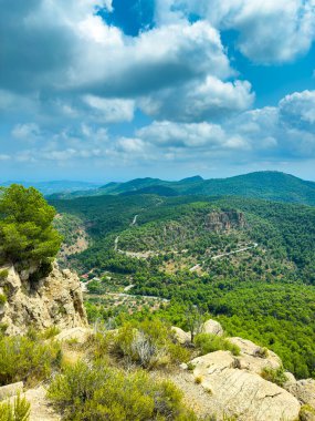 Bereketli yeşil tepeler açık mavi bir gökyüzünün altında yuvarlanıyor ve kayalık kayalıklar manzarayı çerçeveliyor. Parlak öğleden sonra ışığı, dolambaçlı yolları ve canlı yeşillikleri ortaya çıkarıyor, çarpıcı bir doğal manzara yaratıyor..