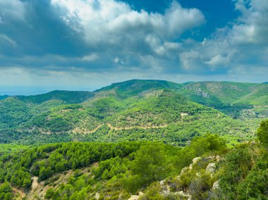 Canlı yeşil tepeler manzara boyunca uzanıyor, dramatik bulutlarla dolu bir gökyüzünün altındaki ağaçlarla benekli. Sahne, keyifli bir günde dışarıdaki aktiviteleri ve keşifleri davet eder..