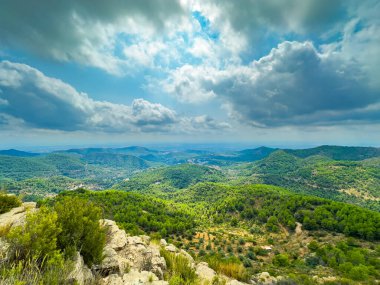 Ağaçlar ve çalılarla kaplı yemyeşil tepeler ufuk boyunca uzanıyor. Gökyüzü bulutlarla dolu, aşağıdaki canlı manzaraya gölgeler düşüyor..