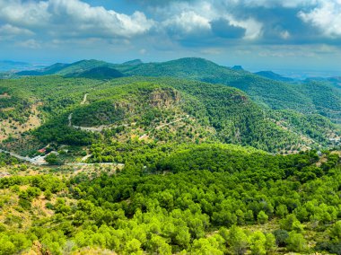 Uçsuz bucaksız bir manzara, bulutlarla benekli mavi gökyüzünün altındaki dağları ve sık ormanları sergiliyor. Güneş yeşilliği aydınlatır, doğanın huzurlu atmosferini güçlendirir..