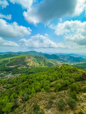 Ufuk boyunca uzanan yemyeşil tepeler ağaçlarla çevrili ve yuvarlanan dağlarla çevrili. Yumuşak bulutlar canlı, mavi bir gökyüzünde süzülür, barışçıl bir atmosfer yaratır..