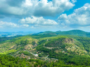 Uçsuz bucaksız yeşil tepeler, kabarık beyaz bulutların altında virajlı yollarla kaplı arazide uzanıyor. Manzara, güneşli bir günde doğanın güzelliğini mükemmel bir uyum içinde sergiliyor..