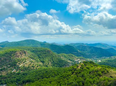 Bereketli yeşilliklerle kaplı geniş tepeler gün ışığıyla aydınlanır ve yükselir. Pofuduk beyaz bulutlar canlı mavi bir gökyüzünde süzülür, doğal güzellik ve sakinliğin resimsel bir sahnesini yaratır..
