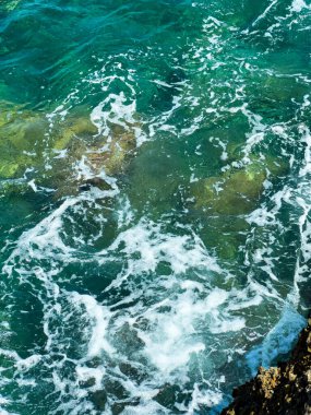 Dalgalar engebeli kayalara çarpıp köpüklü beyaz kabarcıklar oluştururken güneş ışığı berrak turkuaz suda parlıyor. Doğanın ahengi bu kıyı sahnesinde sergileniyor..