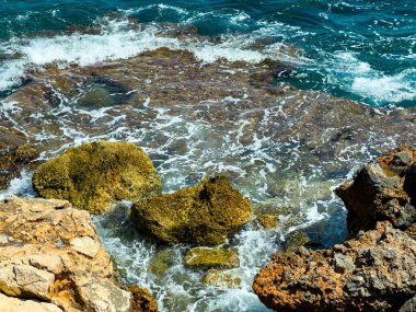 Kıyıdaki engebeli kayalara çarptıklarında parlak güneş ışığı parlak dalgalar üzerinde parlıyor. Gün ortasında sakin bir atmosfer canlı deniz manzarasını çevrelemektedir..