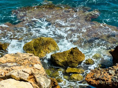 Yumuşak dalgalar engebeli kayaların üzerinde hareket ederken güneş ışığı berrak mavi sularda parıldıyor. Kayaların etrafında deniz köpüğü birikir ve bu sahil çekilmesinin sakin atmosferine katkı sağlar..