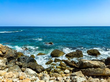 Güneş ışıl ışıl parlarken dalgalar kayalık bir plaja yuvarlanıyor. Açık mavi su ufka doğru uzanıyor ve sakin bir sahil manzarası yaratıyor..