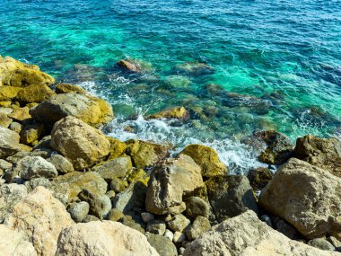 Parlak turkuaz dalgalar kayalık kıyıya hafifçe çarpar, huzurlu bir kıyı gününün özünü yakalar, güneş ışığı suyun yüzeyinde dans eder..