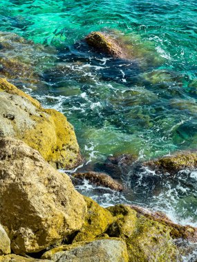 Berrak turkuaz sular engebeli kayalara çarpar ve parlak güneş ışığı altında canlı deniz renkleri sergiler. Dalgalar doğanın sakin sahnesini gözler önüne sererken ritmik olarak dans eder..