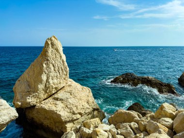 Parlak öğle güneşinin altında, kayalık kıyı şeritleri derin mavi denizden dramatik bir şekilde yükselir. Tekneler ufukta süzülürken kayalara nazikçe vuran dalgalar huzur verici bir manzara yaratıyor..