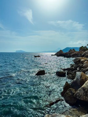 Parlak güneş ışığı sakin denizden yansıyarak su üzerinde ışıl ışıl bir etki yaratır. Küçük bir tekne dalgalı dalgaların arasında süzülürken, Rocky uçurumları kıyı boyunca uzanır..
