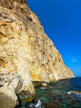 Güneş, denizin yanındaki yüksek ve kayalık bir uçurumda ışıl ışıl parlıyor, yukarıdaki açık mavi gökyüzü ve kıyıya vuran nazik dalgalar, sakin bir kıyı manzarası yaratıyor..