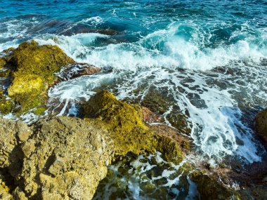 Dalgalar güçlü bir kıyı şeridi boyunca engebeli kayalara çarpar. Okyanus parlak güneş ışığını yansıtır, açık bir günde canlı bir atmosfer yaratır..