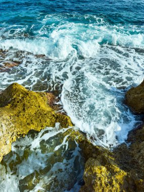 Parlak mavi dalgalar, sakin bir sahilde altın kayalara çarpar. Gün ortası güneşi altında köpüklenen ve parıldayan köpüklü su, canlı bir kıyı sahnesi yaratır..