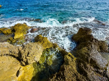 Dalgalar engebeli kayalara hafifçe çarparken suyun üzerinde parlak güneş ışığı parıldıyor. Mavi deniz, kıyı şeridinin topraksı tonlarıyla çok güzel zıtlaşır ve sakin bir kıyı manzarası yaratır..