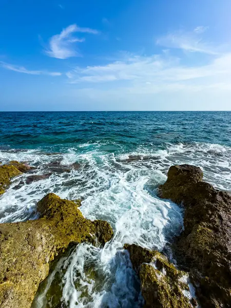 Berrak turkuaz sular engebeli kayaların üzerinde yavaşça yuvarlanarak güzel bir kontrast oluşturuyor. Güneş parıldıyor, bu kıyı cennetinin huzurlu atmosferini güçlendiriyor..