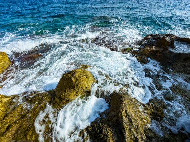 Parlak güneş ışığı, dalgaların engebeli kayalara çarpmasıyla sulardan yansıyarak, sahilin dingin güzelliğini vurgulayan köpük ve sıçramalar yaratarak, keşif ve barışı davet eder..