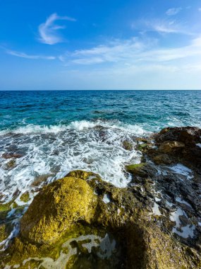 Dalgalar kıyı şeridi boyunca uzanan engebeli kayalara hafifçe çarparak güneş altında parlar. Açık mavi bir gökyüzü birkaç kabarık bulutla birlikte uzanıyor ve sakin bir atmosfer yaratıyor..