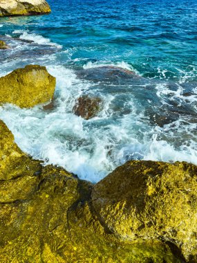 Dalgalar canlı bir kıyı şeridi boyunca engebeli kayalara karşı enerjik bir şekilde dalgalanır. Su güneşin altında parlıyor, doğanın güzellikleriyle dolu canlı bir açık hava manzarası yaratıyor..