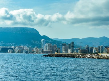 Açık mavi sulara bakan ziyaretçiler, bulutlu bir günde arka planda yükselen dağlarla kıyı şeridi boyunca uzanan modern binaları görebilirler..