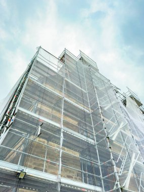 Koruma ağlarına sarılı yüksek bir bina iskele arasında duruyor ve yoğun inşaat sürecini ortaya çıkarıyor. Bulutlu gökyüzü, sahneye dramatik bir dokunuş ekler..