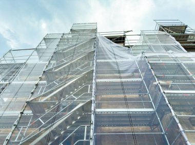 İnşaat halinde olan ve parlak bir günde koruyucu ağlarla örülmüş yüksek bir binanın etrafını iskeleler sarar. İşçiler yerin üstünde malzeme hazırlarken görülebilir..