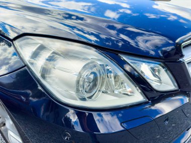 Göz kamaştırıcı mavi bir araba güneş ışığında parıldıyor. Detaylı farlarını ve parlak dış görünüşünü sergiliyor. Kabarık bulutlar yukarıdaki parlak gökyüzünde sakin bir şekilde süzülür ve manzaraya güzellik katar..