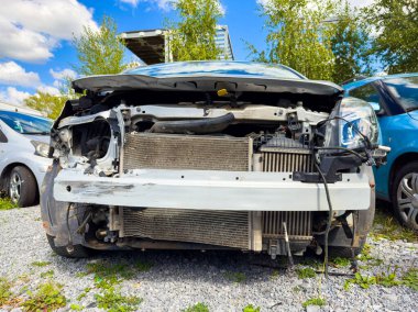 Hasarlı bir araba bir hurdalıkta ön tarafı açıkta ve önemli derecede ön tarafı hasar görmüş bir şekilde durur. Diğer araçlar arka planda açık mavi gökyüzünün altında görülür..