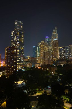 Gökdelenler karanlık bir gökyüzüne karşı parıldıyor, pencereleri sıcak ışıklarla parıldıyor. Aşağıdaki ağaçlar ve sokaklar bu hareketli şehir ortamını rahatlatıyor..