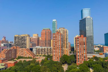 Şehir silueti, yeşil parklar ve ağaçlarla çevrili modern gökdelenler ve zarif konutların karışımından oluşan açık mavi gökyüzü altında enerji yayıyor ve canlı bir şehir manzarası yaratıyor..