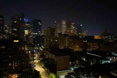 Canlı bir şehir silueti gece gökyüzüne karşı parlak bir şekilde parlıyor. Yüksek binalar parlak ışıkları yansıtırken, sessiz sokaklar şehrin canlı atmosferine ipucu veriyor..