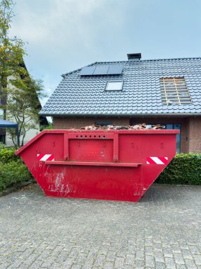 Canlı kırmızı bir çöp bidonu, kayıp çatı kiremitleri ile görülebilir onarımları olan tuğla bir evin önünde belirgin bir şekilde duruyor. Sakin bir muhitte, ev gelişimini vurgulayan açık bir gün..