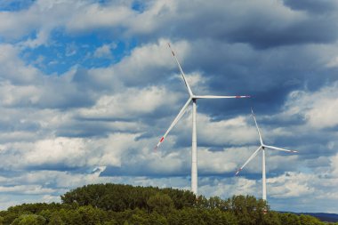 İki uzun rüzgar türbini, yumuşak, beyaz bulutlarla dolu engin bir gökyüzünün altındaki yeşil bir tepede yavaşça dönüyor. Sahne, doğa ve temiz enerjinin barışçıl birlikteliğini yakalar..