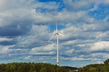 Bereketli yeşil orman üzerinde güçlü bir rüzgar türbini, dinamik bulutlarla dolu geniş bir gökyüzü ile çevrili, barışçıl bir kırsal alanda yenilenebilir enerji sergiliyor..