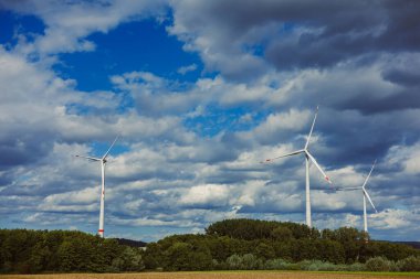 Üç rüzgar türbini mavi bir gökyüzü altında zarif bir şekilde dönüyor. Etrafı canlı yeşil tarlalarla çevrili bulutlarla kaplı. Bu sahne sürdürülebilir enerji kaynaklarının güzelliğini vurgular..
