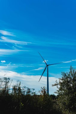 Uzun bir rüzgar türbini yeşilliğin üzerinde gururla yükselir, rüzgarlı bir öğleden sonra pervaneleri yavaşça döner. Gökyüzü ince bulutlarla dolu ve parlak mavi bir renk, doğanın güzelliğini gözler önüne seriyor.
