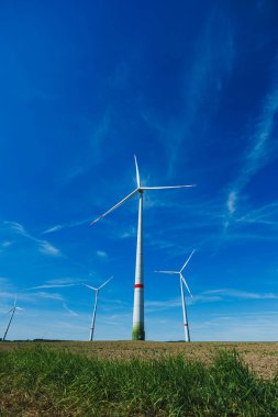 Barışçıl bir kırsal bölgedeki açık mavi gökyüzünün arka planında belirgin bir şekilde yükselen üç rüzgar türbini, sürdürülebilir enerjiyi eylem halinde gösteriyor.