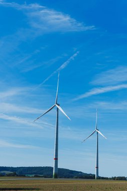 Uzun rüzgar türbinleri, güneşli bir günde kırsal alanlarda temiz enerji kullanımını işaret eden, barışçıl yeşil alanlarla çevrili, canlı mavi gökyüzüne karşı zarifçe döner..