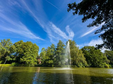 İlk ışıkta, bir çeşme suyu zarif bir şekilde canlı yeşil ağaçlarla çevrili bir göle püskürtürken ince bulutlar yukarıdaki mavi gökyüzünde tembelce sürükleniyor..