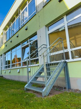 Metal bir merdiven çimenli bölgeyi modern yeşil bir binaya bağlar. Gün ışığı parlak adımları yansıtır, gün boyunca davetkar girişi vurgular..