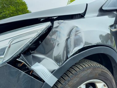 Siyah bir araç ön yolcu tarafında ciddi hasar gösteriyor. Metalde göçükler ve yırtıklar var. Bulutlu gökyüzü yemyeşil manzaranın yanında kasvetli bir hava katıyor..