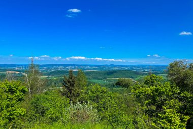 Sahne, parlak ve bulutsuz bir gökyüzü altında yeşil vadiler ve tepeler gösteren, yüksek bir bakış açısından çarpıcı bir manzara yakalıyor. Doğa her yerde gelişir, ağaçlar ön planda dağılır..
