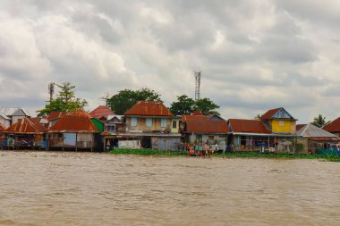 Palembang, 22 Aralık 2024: Bulutlu gökyüzü altında eşsiz bir mimari ve yemyeşil bitki örtüsüne sahip nehir kenarı köyü.