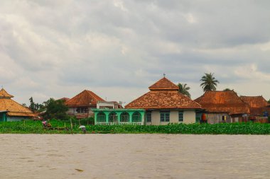 Palembang, 22 Aralık 2024: Bulutlu gökyüzü altında eşsiz bir mimari ve yemyeşil bitki örtüsüne sahip nehir kenarı köyü.