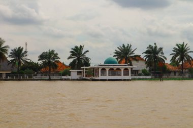 Palembang, 22 Aralık 2024: Bulutlu gökyüzü altında eşsiz bir mimari ve yemyeşil bitki örtüsüne sahip nehir kenarı köyü.