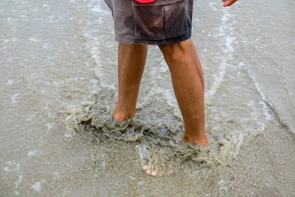Sığ sularda yürüyen bir çocuğun bacakları kumlu bir sahilde neşe saçar..