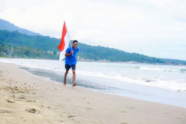 Endonezyalı adam kumlu sahil şeridi boyunca bayrağıyla neşeyle koşuyor..