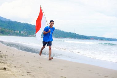 Adam Endonezya bayrağıyla kumlu bir sahil boyunca koşuyor, Bağımsızlık Günü 'nü kutluyor.