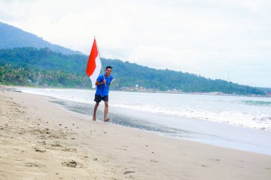 Genç Endonezyalı bir adam dingin bir sahilde yürürken kırmızı ve beyaz bayrak sallıyor.